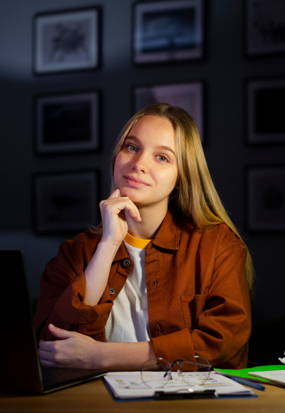 Junge Frau sitzt am Schreibtisch und blickt selbstbewusst in die Kamera, während sie ihre Hand am Kinn hält. Im Hintergrund sind gerahmte Bilder an der Wand zu sehen. Moderne Lernumgebung und Konzentration bei der Arbeit im Homeoffice.