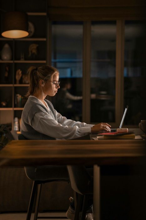 Junge Frau arbeitet konzentriert am Laptop in einem modernen, gemütlich beleuchteten Raum. Sie sitzt an einem Schreibtisch mit Regalen im Hintergrund, die eine ruhige Lernumgebung im Homeoffice darstellen.
