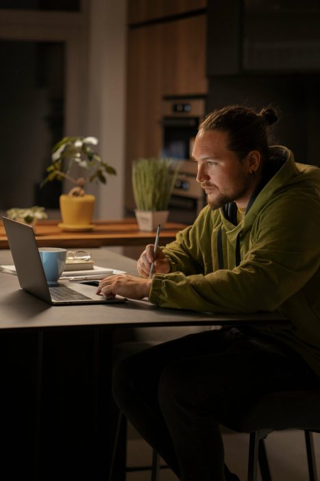 Junger Mann arbeitet konzentriert am Laptop in einer modernen Küche, während er Notizen macht. Die ruhige und stilvolle Umgebung unterstützt das effektive Lernen und Arbeiten im Homeoffice.